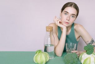 A young woman in a green top sitting with vegetables and a glass of water.